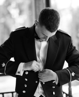 A groom in a formal black tuxedo adjusts his cufflink in an elegant black and white photograph.
