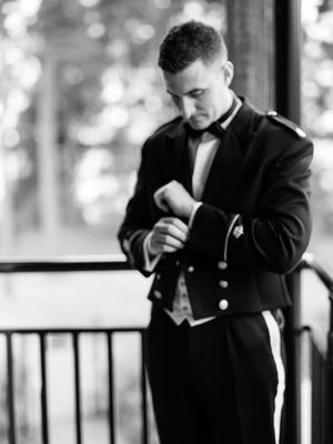 A groom in a formal black tuxedo and bow tie adjusts his sleeve cuffs on an outdoor porch.