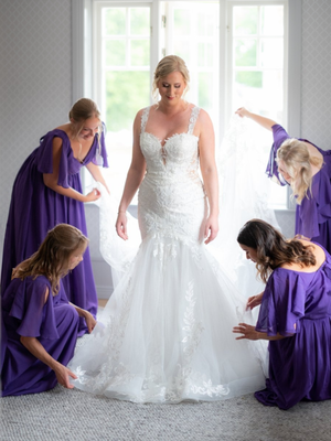 A woman in a white wedding dress kneels next to another in a purple dress against a light-filled interior backdrop.