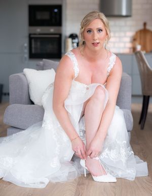 A woman in a flowing white wedding dress sits elegantly on a modern grey sofa while adjusting her shoes.