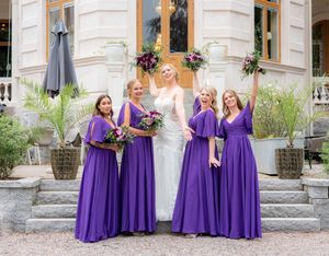A bridal party poses on stone steps wearing matching purple floor-length bridesmaid dresses and holding floral bouquets.