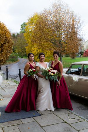 Bridesmaids in burgundy gowns and bride in white dress pose together on autumn day with vintage car in background.