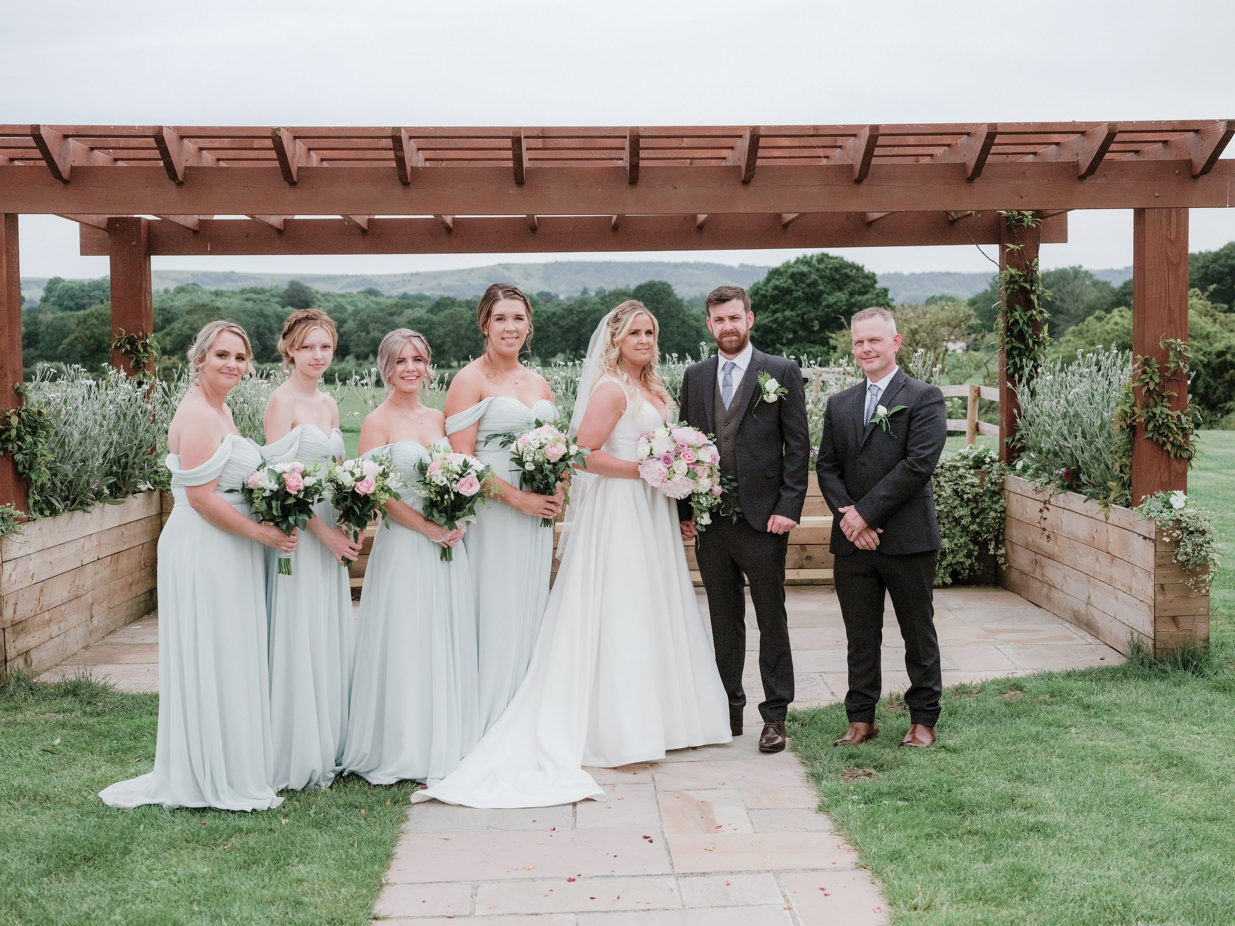 A wedding party poses under a wooden pergola in a garden setting with mint green bridesmaids dresses and black suits.