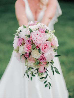 A romantic bridal bouquet with pink and white roses, peonies and greenery against a white wedding dress and grass background.