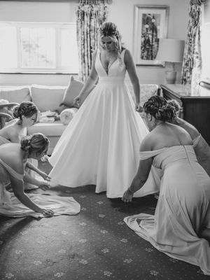 Black and white photo showing bridesmaids helping adjust a flowing white wedding dress in an elegant room.