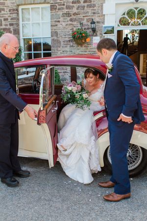 Wedding party helping bride into vintage cream and burgundy classic car outside stone building.