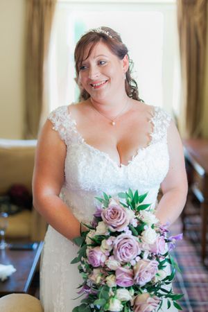 A bride in a white lace wedding dress smiles while holding a beautiful bouquet of pink roses and greenery.