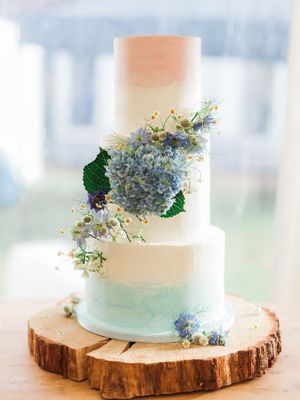 Three-tiered wedding cake with peach and mint ombre design decorated with blue hydrangeas on a rustic wood slice.