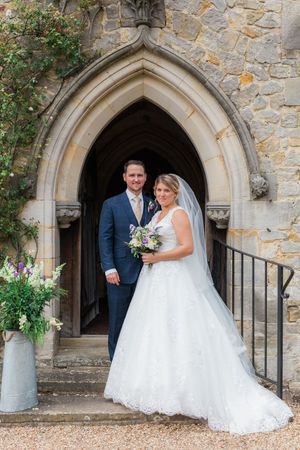 A bride in a white lace gown and groom in navy suit pose under a gothic stone church archway with a floral bouquet.