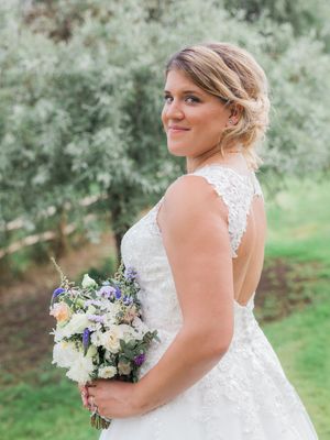 A bride in a lace wedding dress holds a romantic bouquet of white and purple flowers in an outdoor garden setting.