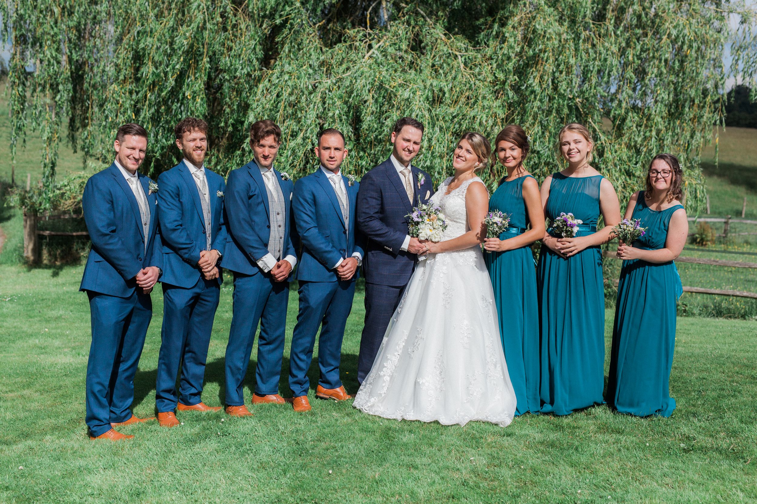 Wedding party in navy blue suits and teal bridesmaid dresses posing together on a sunny green lawn.