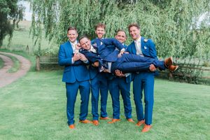 Groomsmen in blue suits sit casually on a grassy lawn under a weeping willow tree.