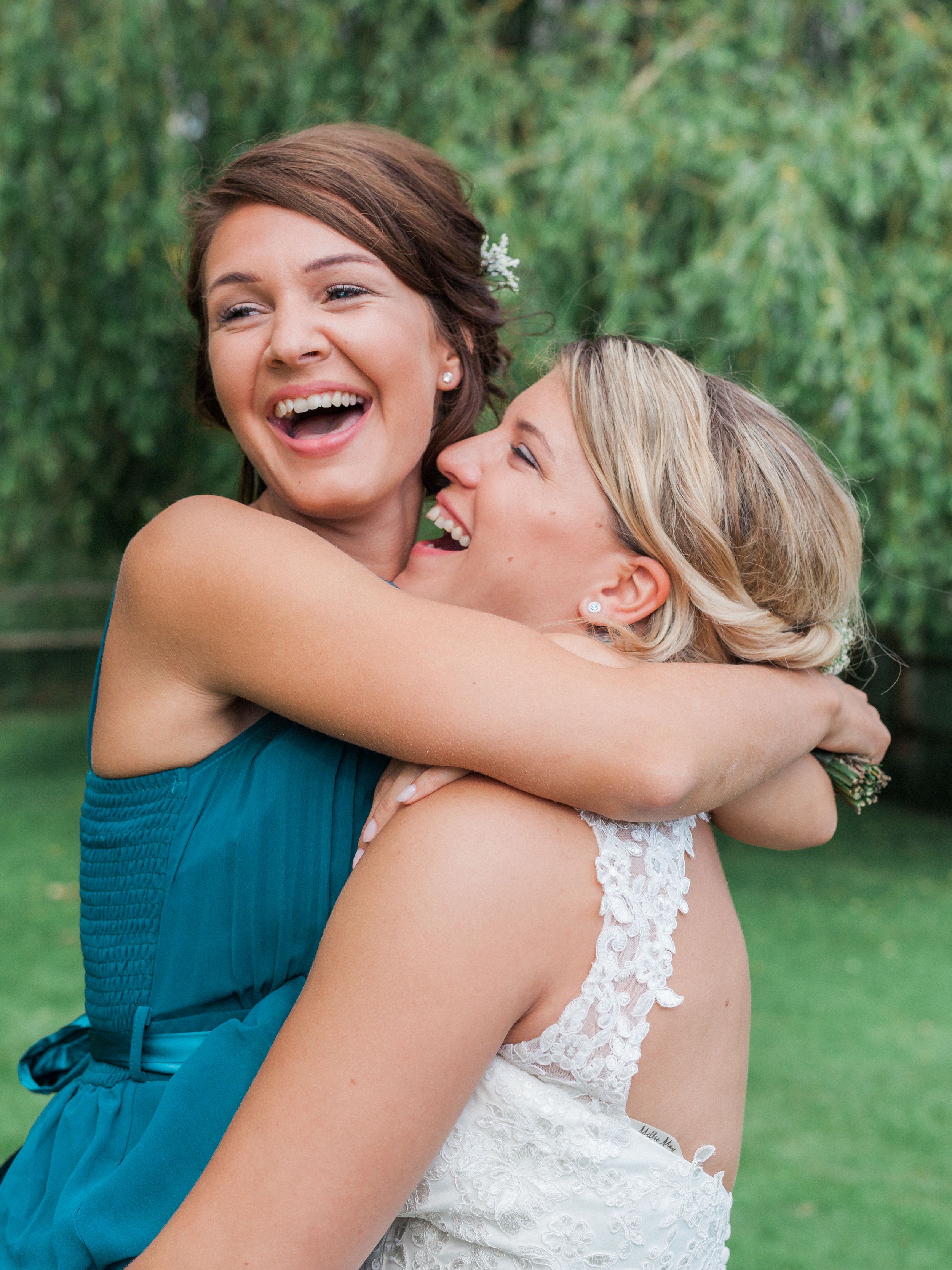 Wedding party members share joyful embraces on a lush green lawn with trees in the background.