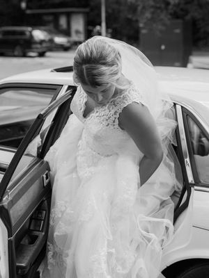 A bride wearing a lace wedding dress with a flowing veil steps elegantly into a white car on her wedding day.