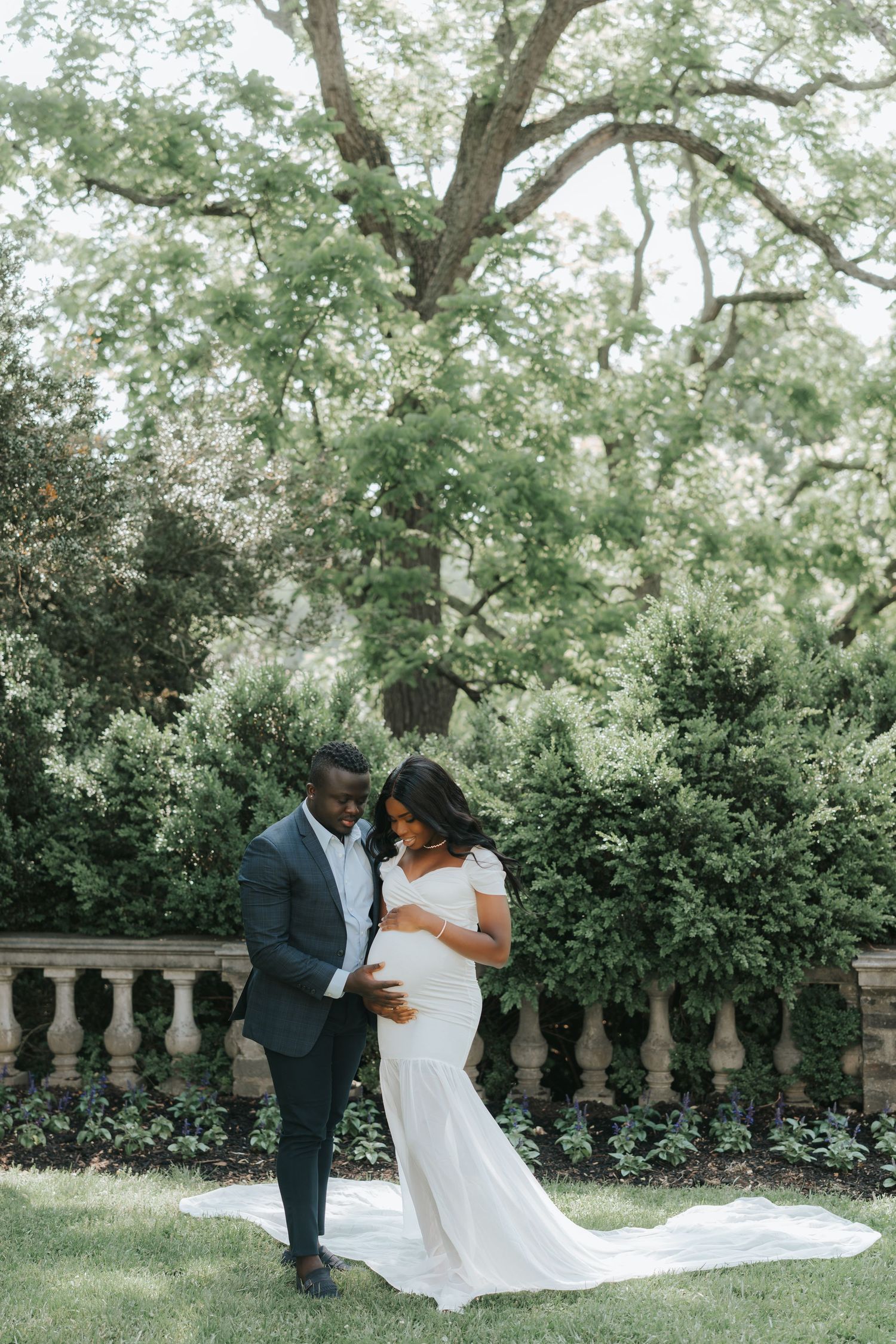 A couple in formal wedding attire embraces in a lush garden setting with stone balustrades and tall trees in the background.