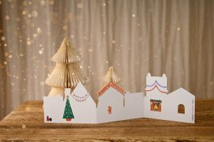 Decorative paper winter village scene with folded trees and white houses against sparkly backdrop.