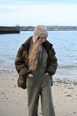 Winter fashion photo showing olive green overalls and brown puffer jacket against coastal backdrop.
