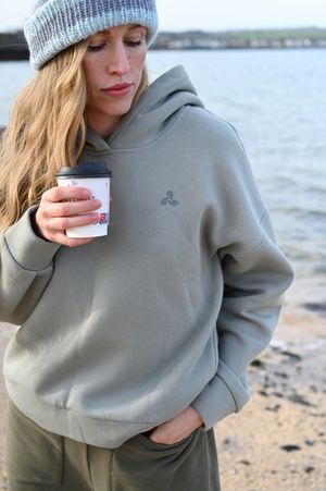 Casual seaside portrait in grey hoodie holding coffee cup on rocky beach.