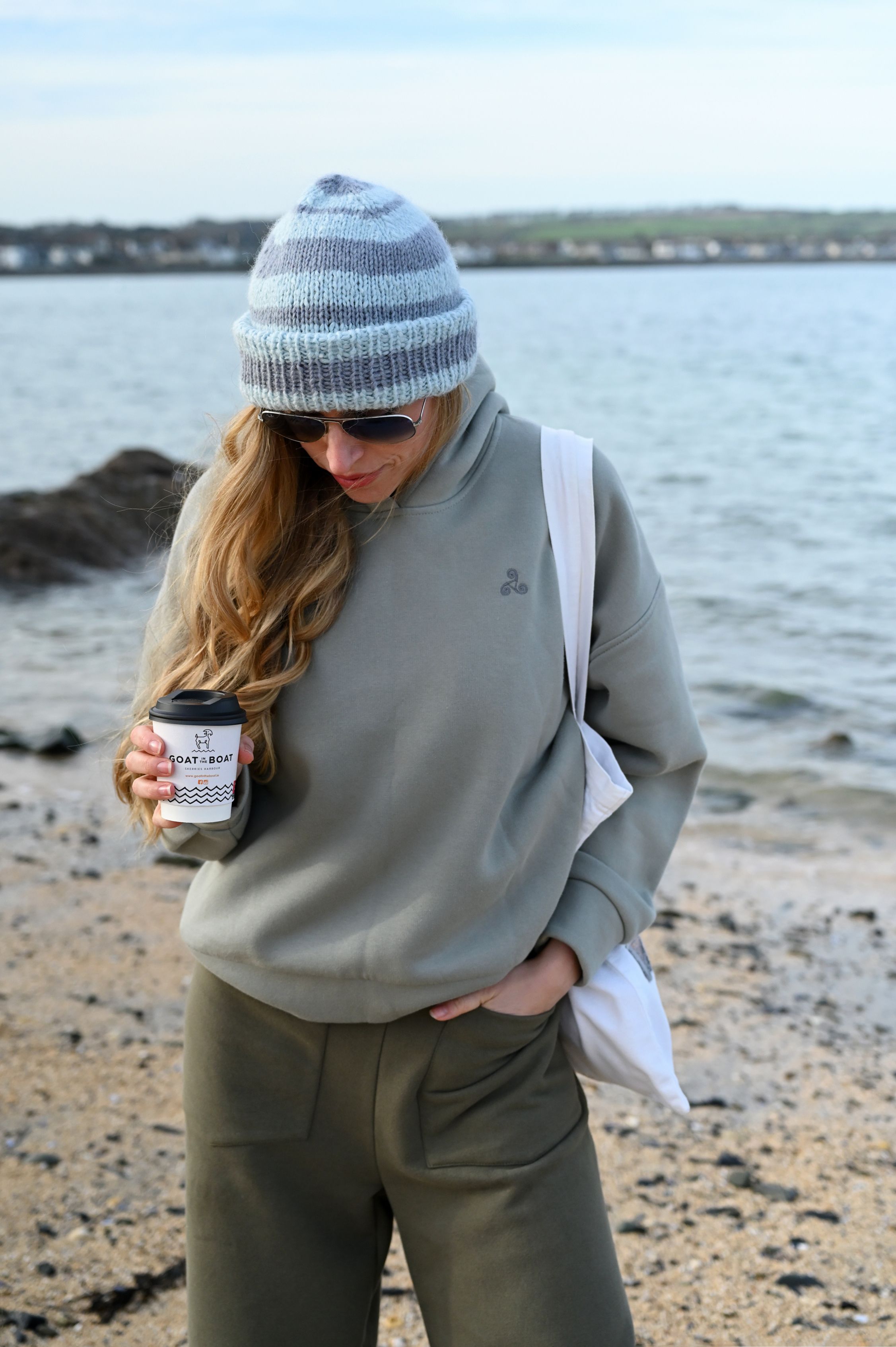 Beachside scene with cozy grey sweatshirt and striped beanie while holding coffee.