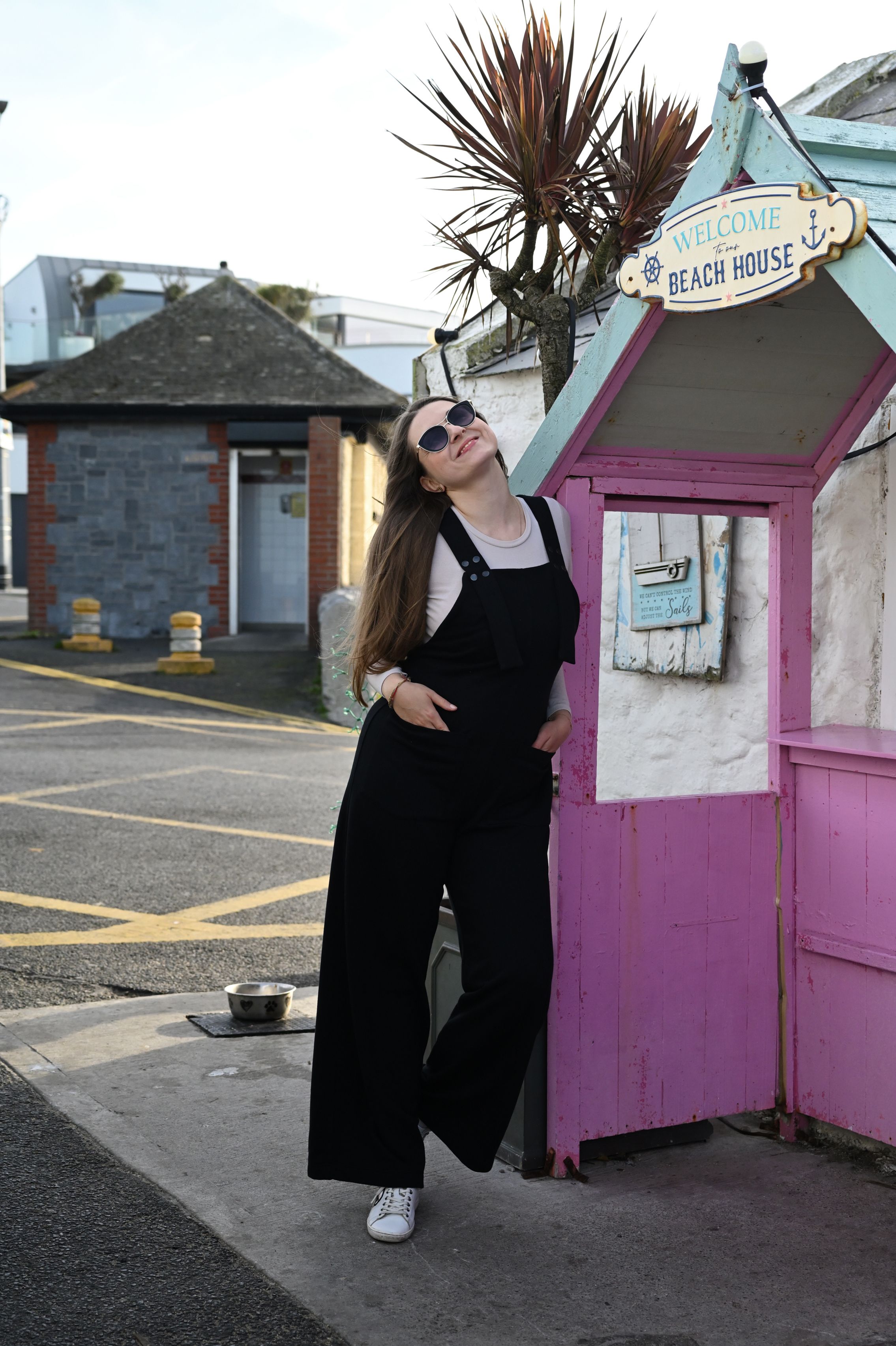 Casual pose by a pink beach house kiosk wearing black overalls and sunglasses.