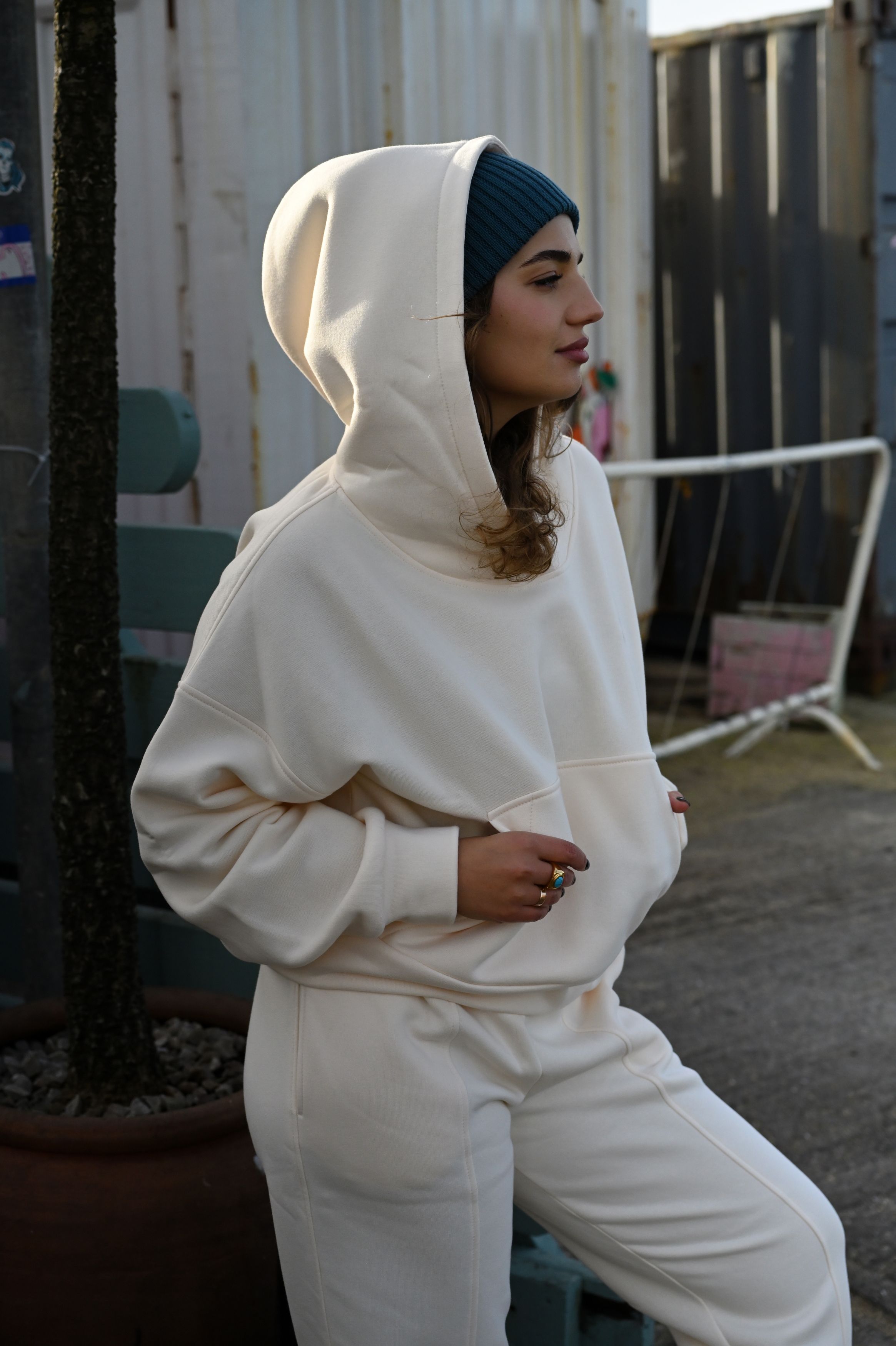 Person in white matching hoodie and sweatpants set with blue beanie poses outdoors.
