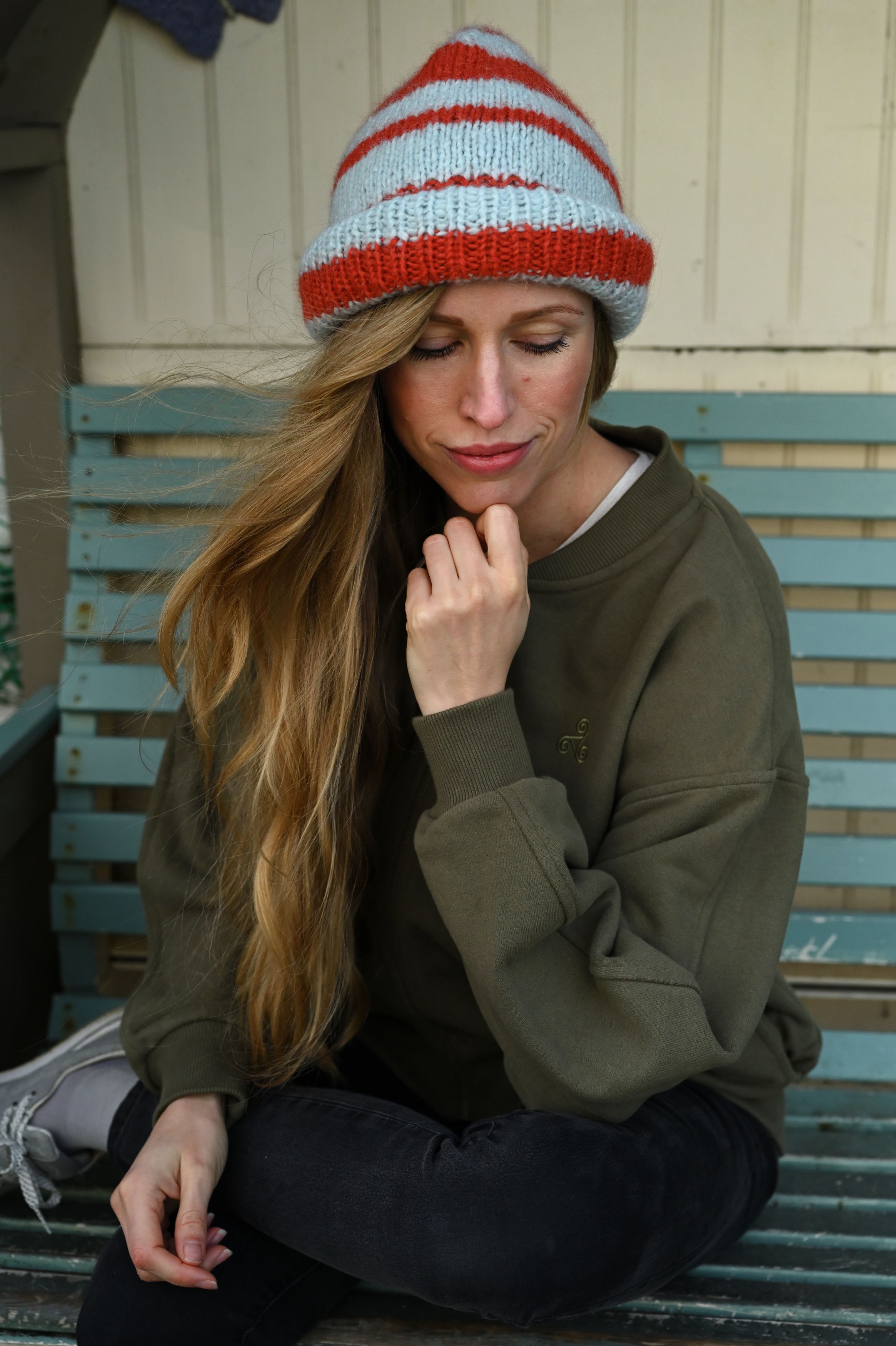 Person in olive sweatshirt and striped beanie sitting thoughtfully on wooden bench.