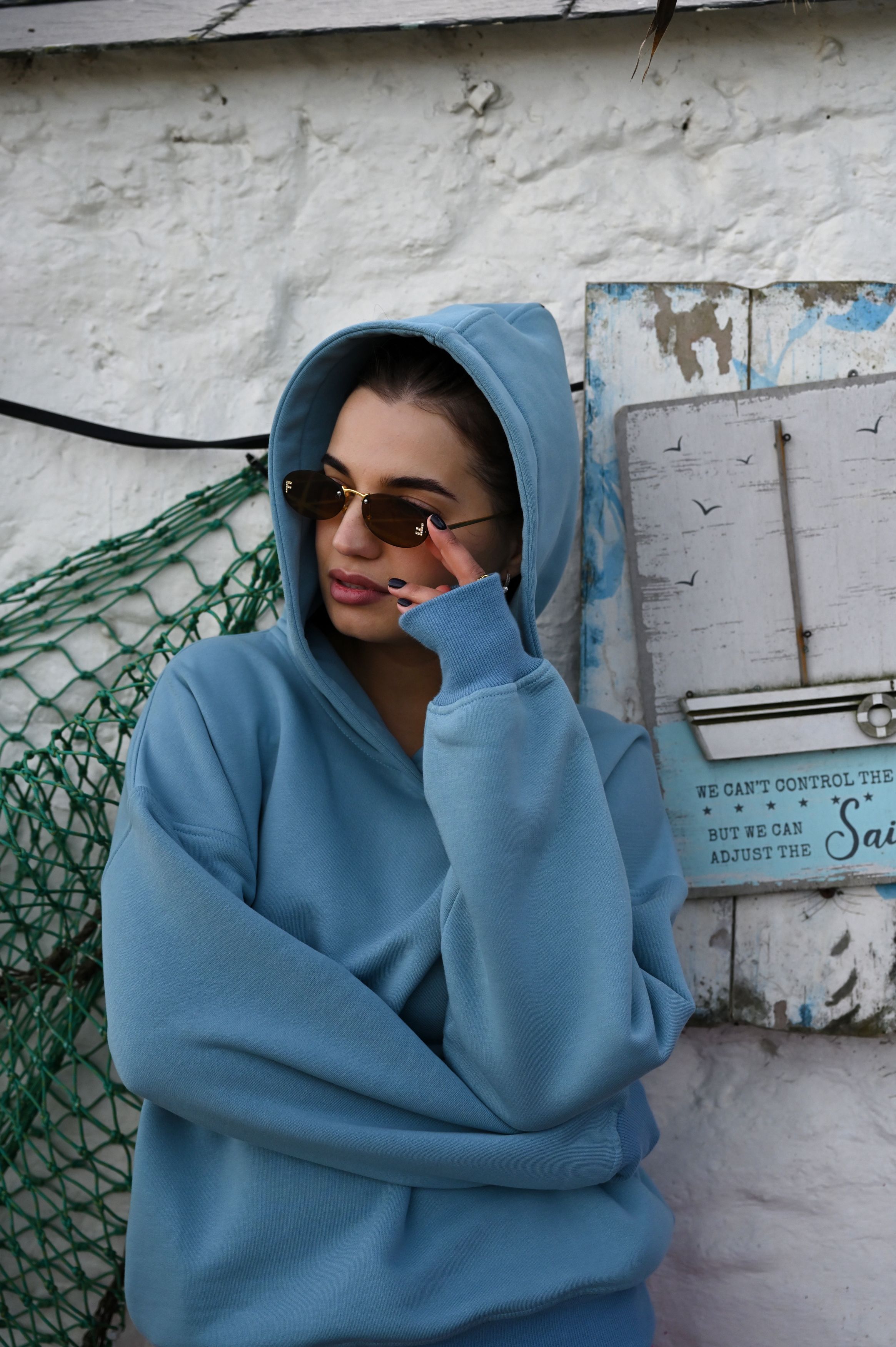 Person in blue hoodie and sunglasses posing against white wall with fishing net backdrop.