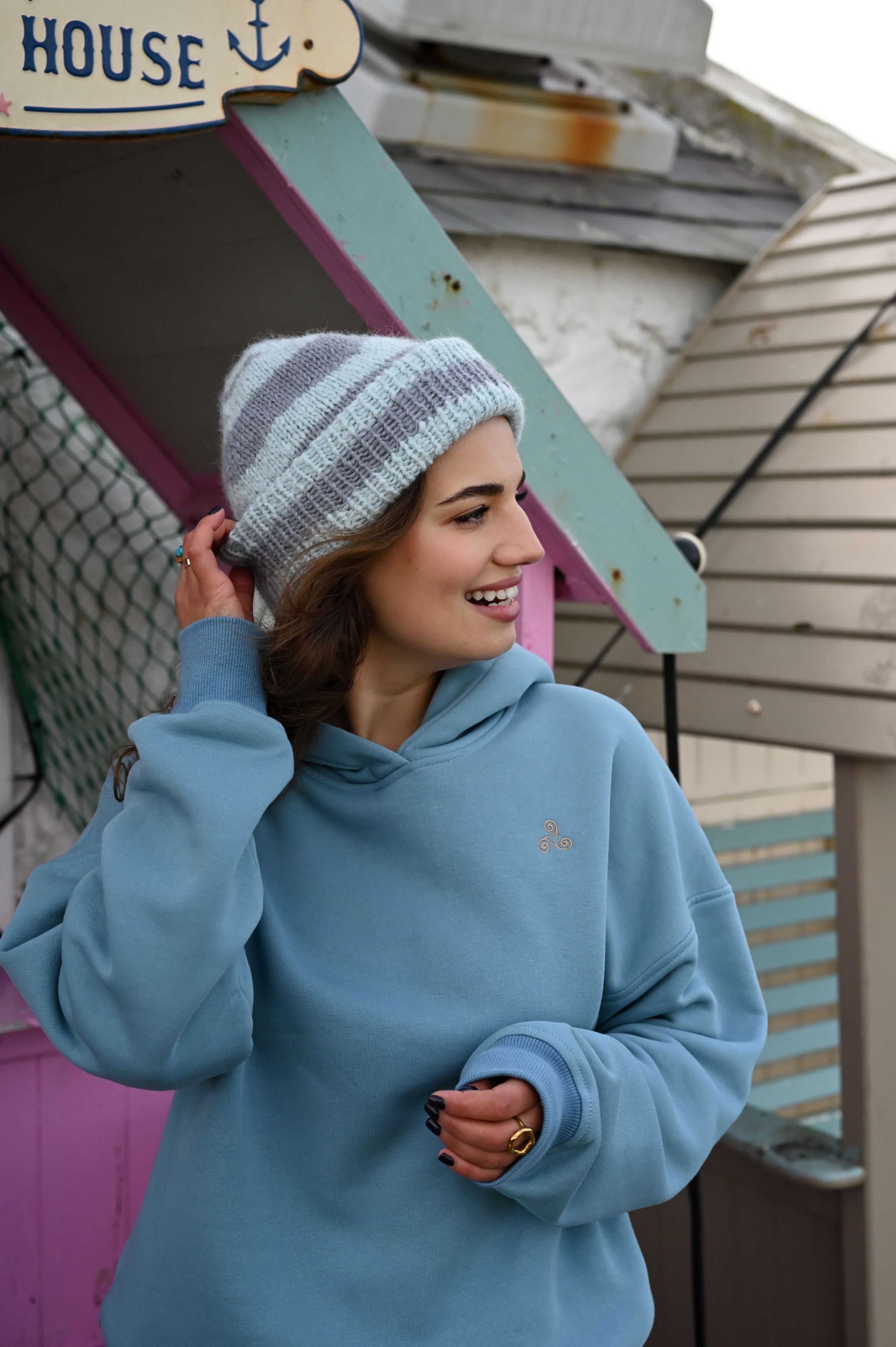 Person in light blue oversized hoodie and beanie smiling near nautical-themed building.