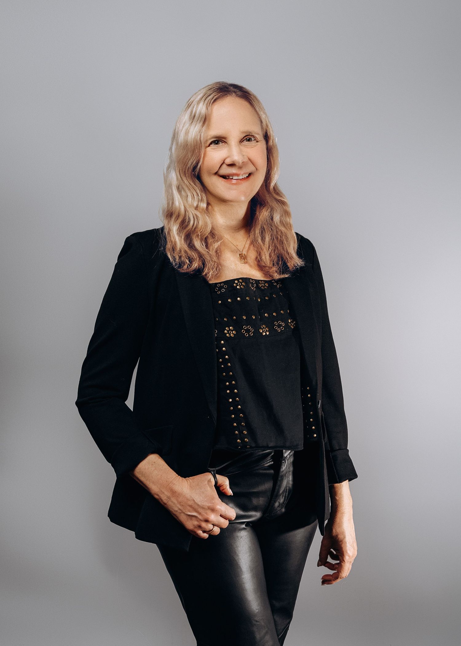 Woman in black blazer and leather pants smiling confidently against a neutral gray studio background.