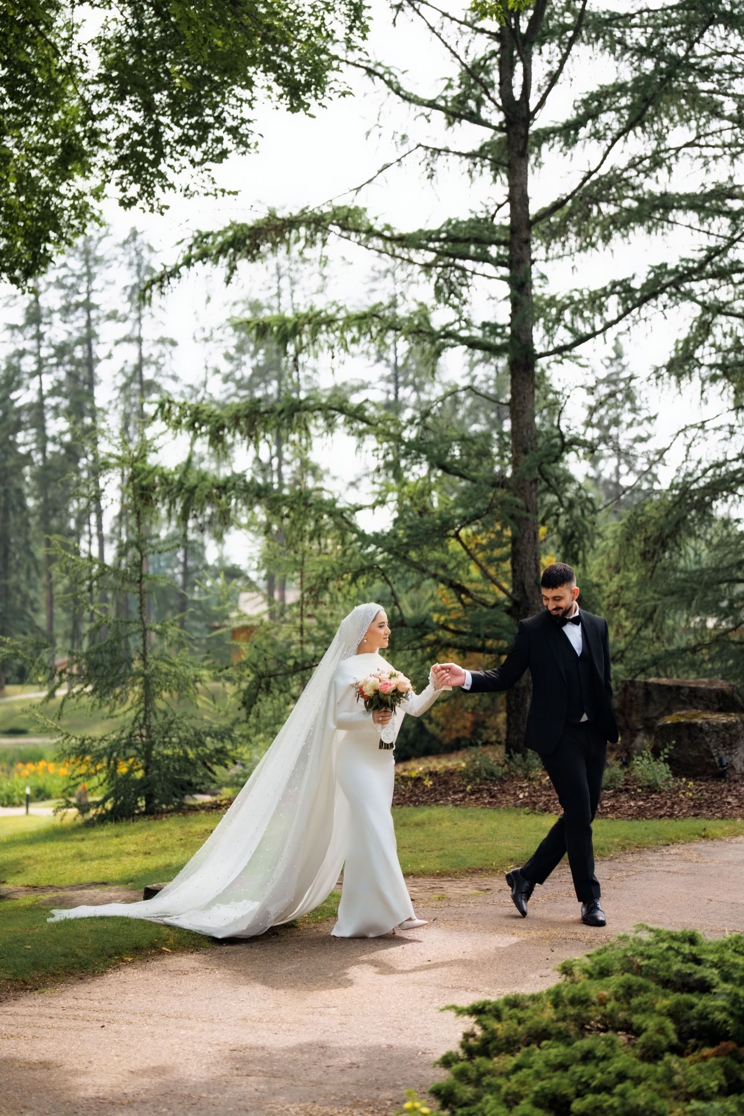 Bride in an off-shoulder lace wedding gown holds pink bouquet near elegant garden fountain on sunny day.