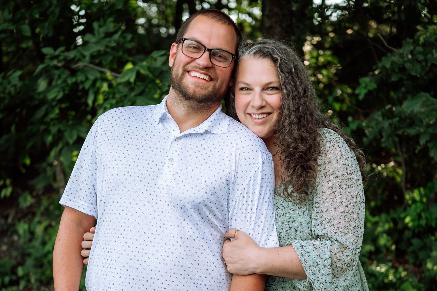 A couple in casual summer attire smiles together in front of leafy green foliage.