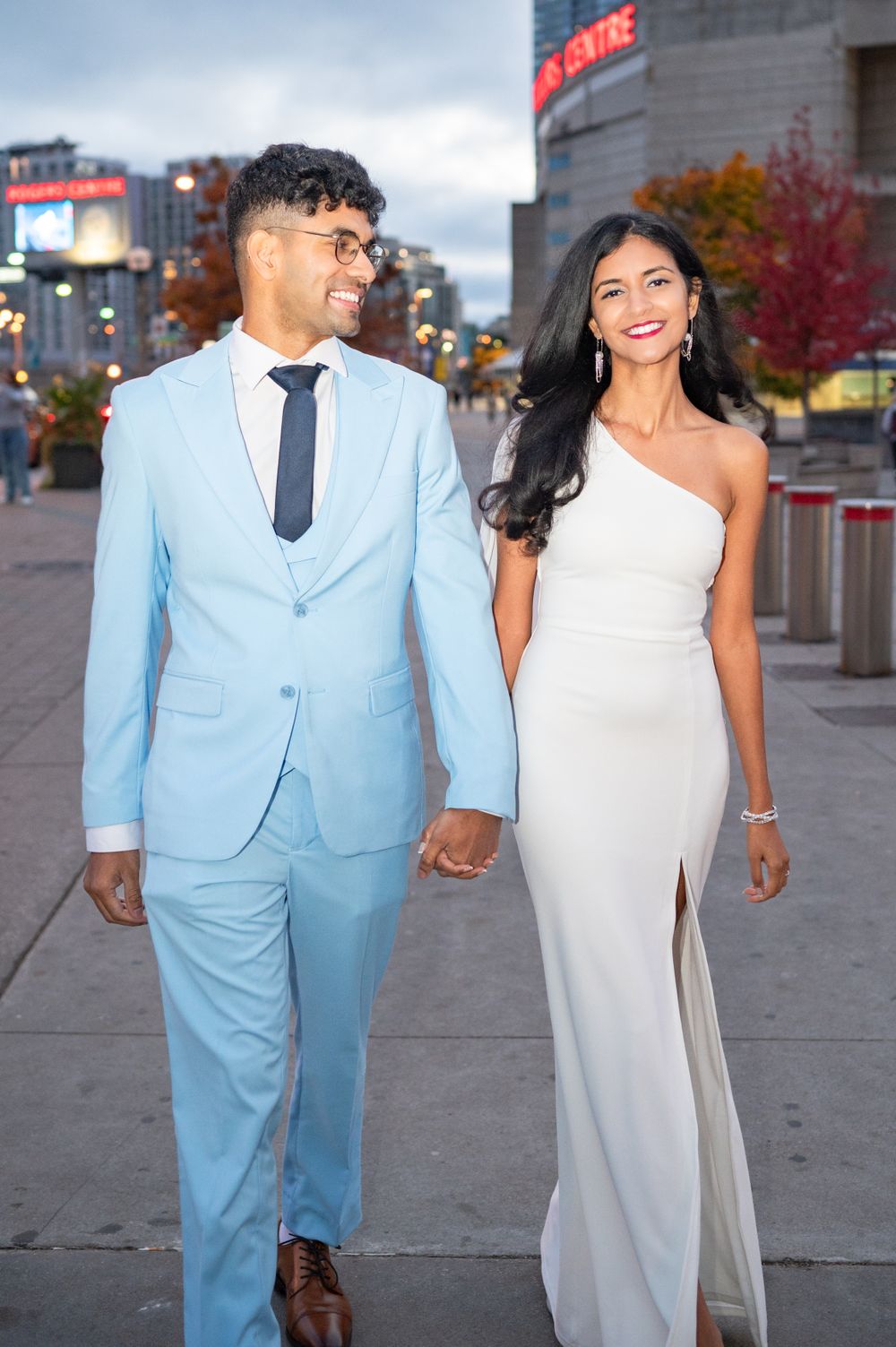 A couple in formal attire walks together outside, with him wearing a light blue suit and her in an elegant white gown.