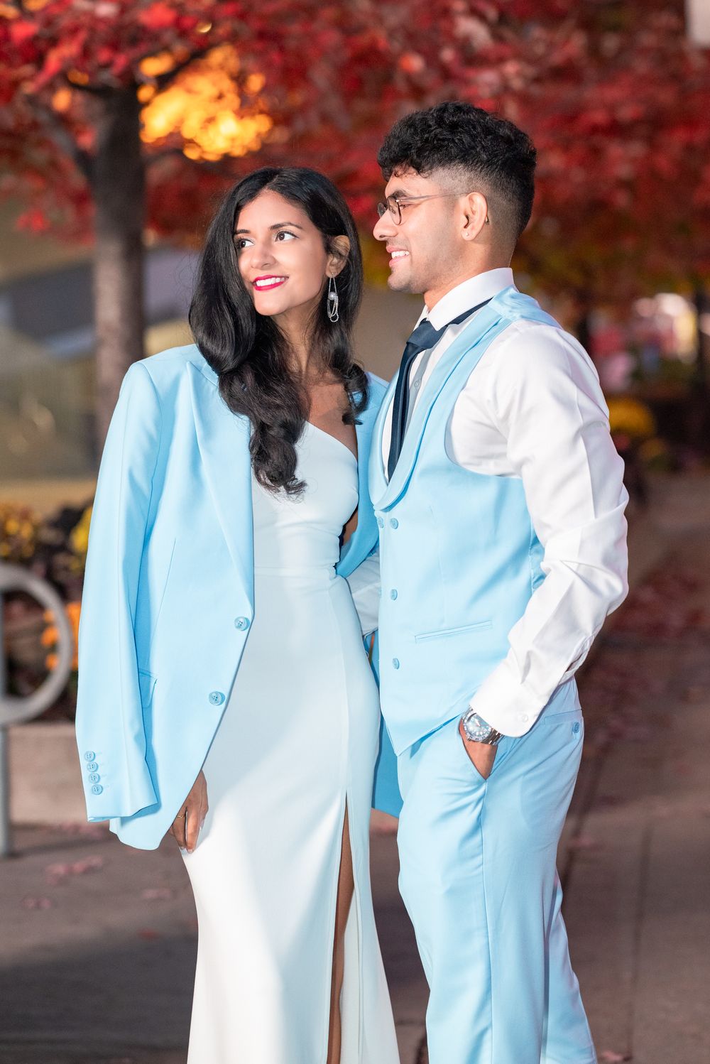 A couple in coordinated light blue and white formal attire pose together against a backdrop of autumn red foliage.