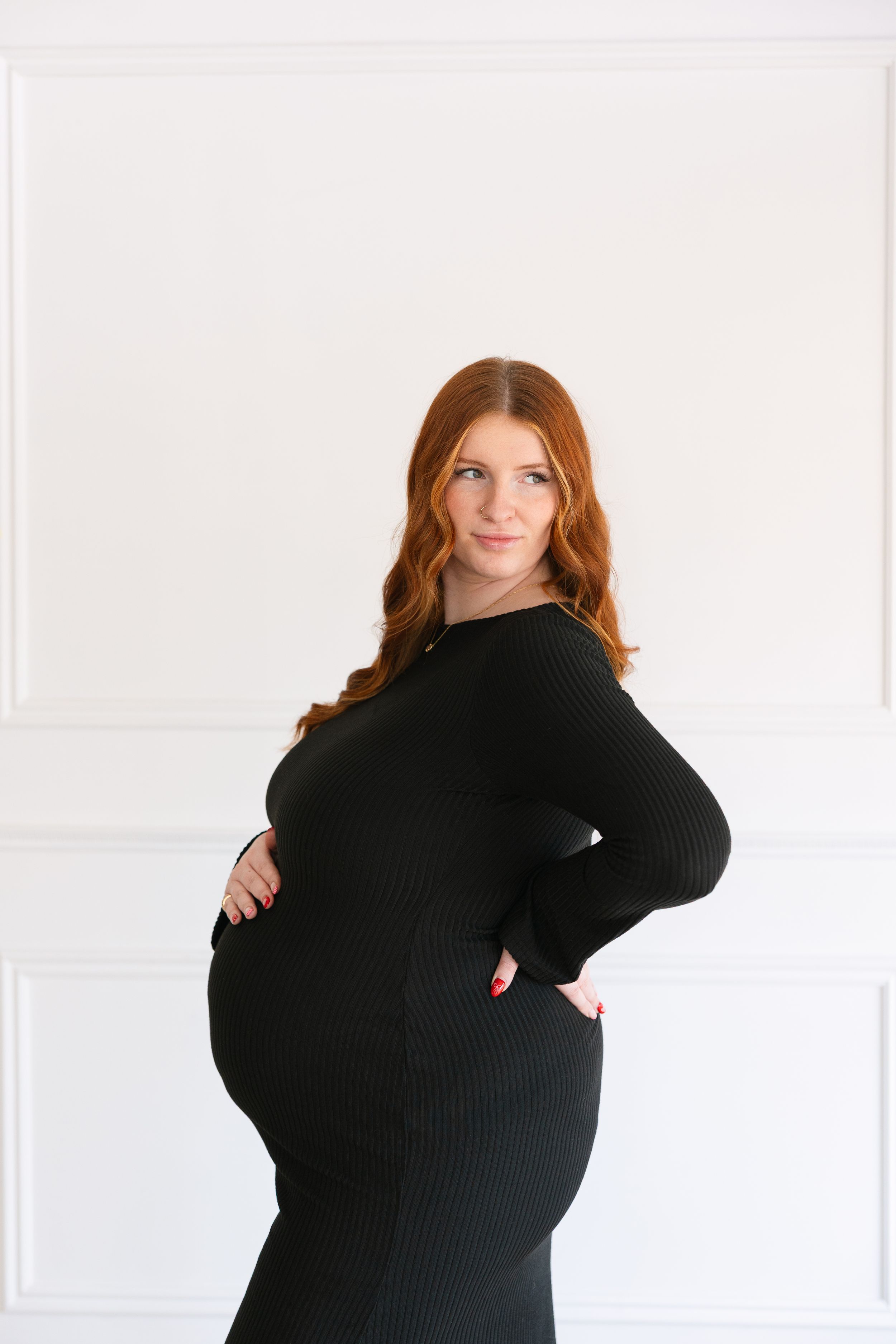 Pregnant woman in black ribbed dress with red hair posing sideways, showing baby bump against white wall.