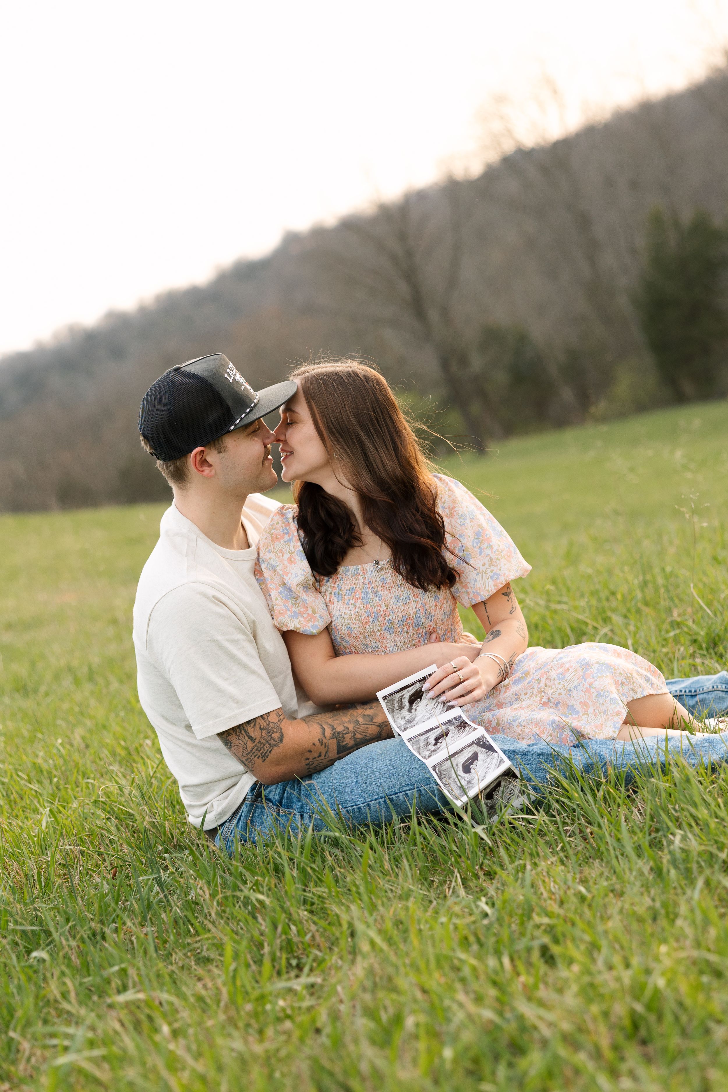 Couple sitting in green field kissing while holding ultrasound photos, announcing pregnancy outdoors.