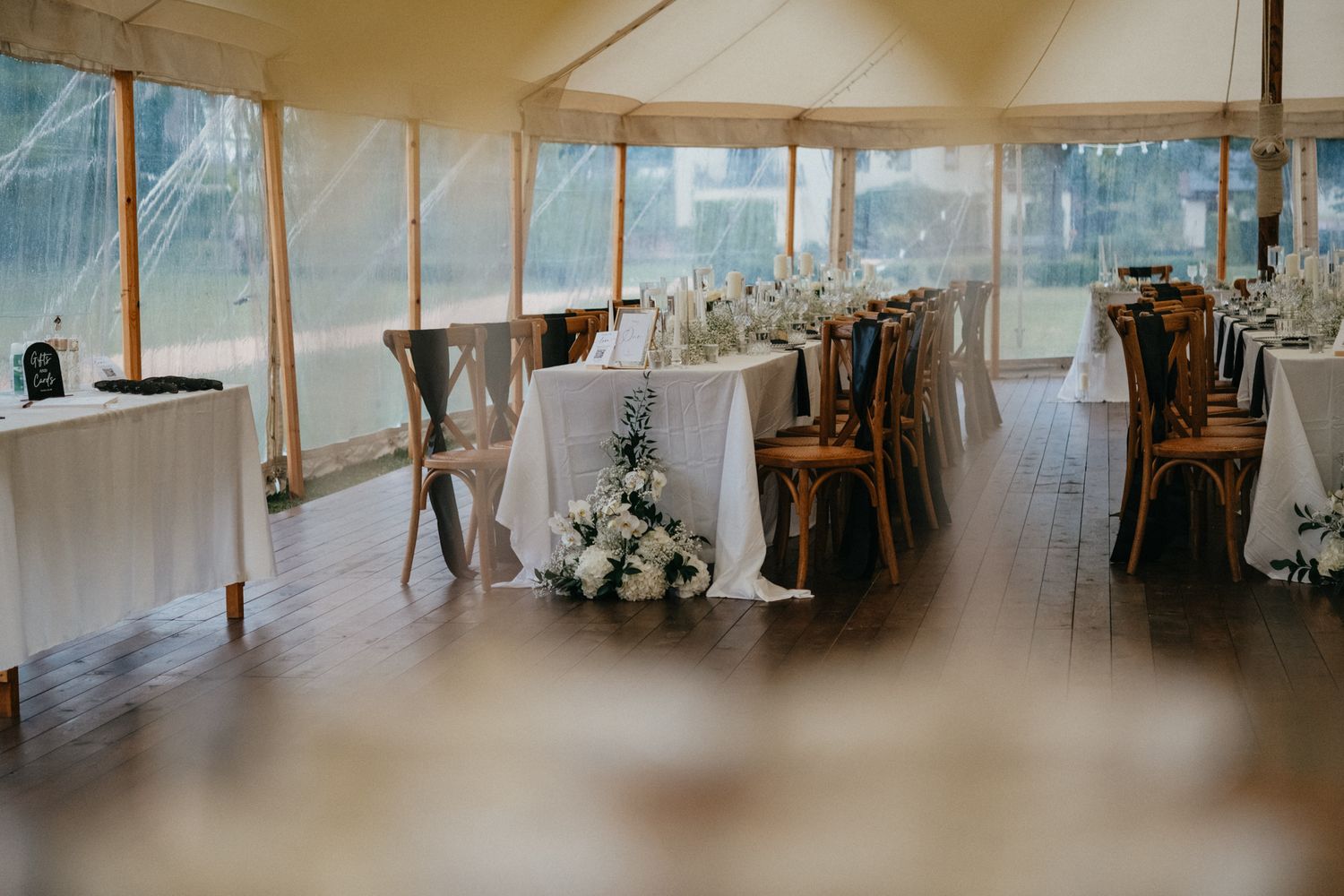 Wedding reception setup inside an elegant tent with wooden crossback chairs and white-clothed tables with flower centerpieces.