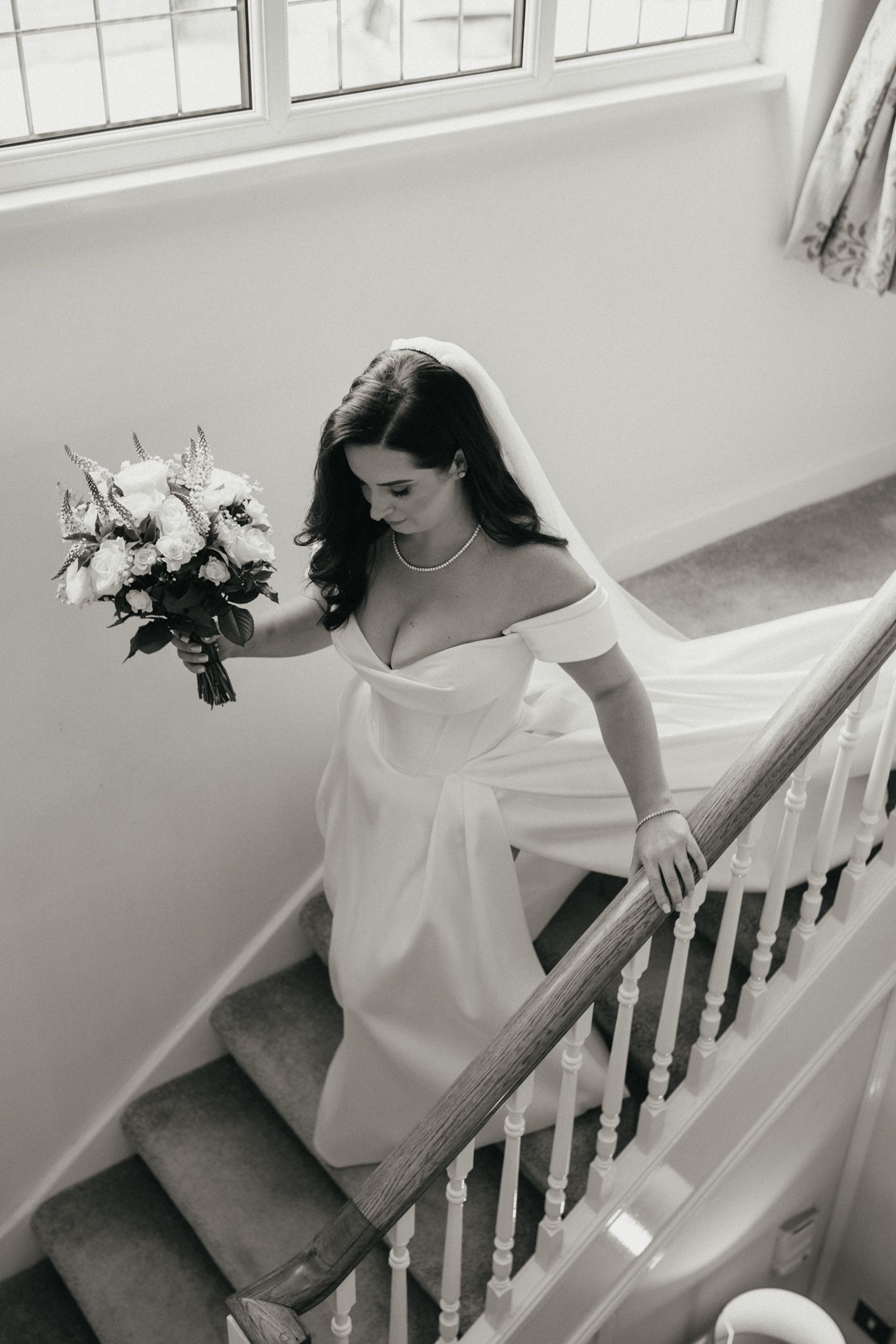 A bride in an elegant off-shoulder gown descends a white staircase while holding a bouquet.