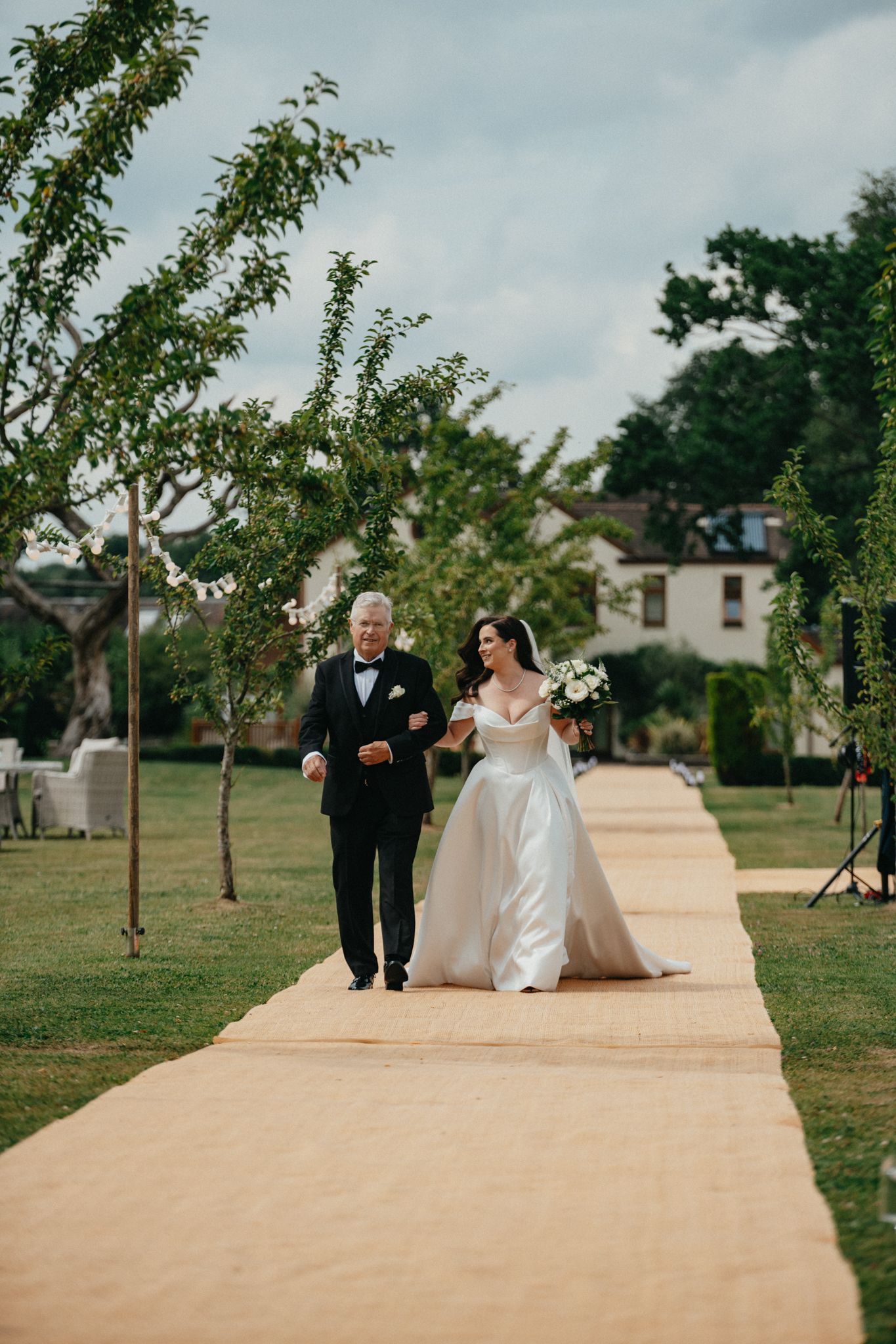 An elegant wedding processional along a beige carpet pathway lined with young trees.