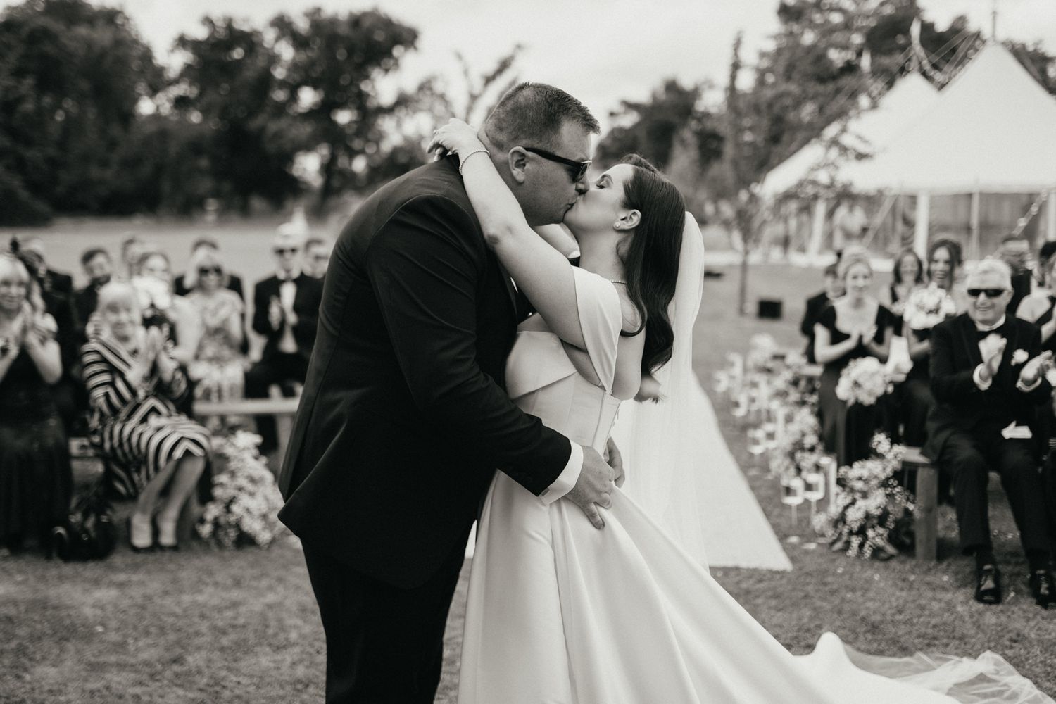 A romantic wedding ceremony kiss captured in black and white with guests watching in the background.