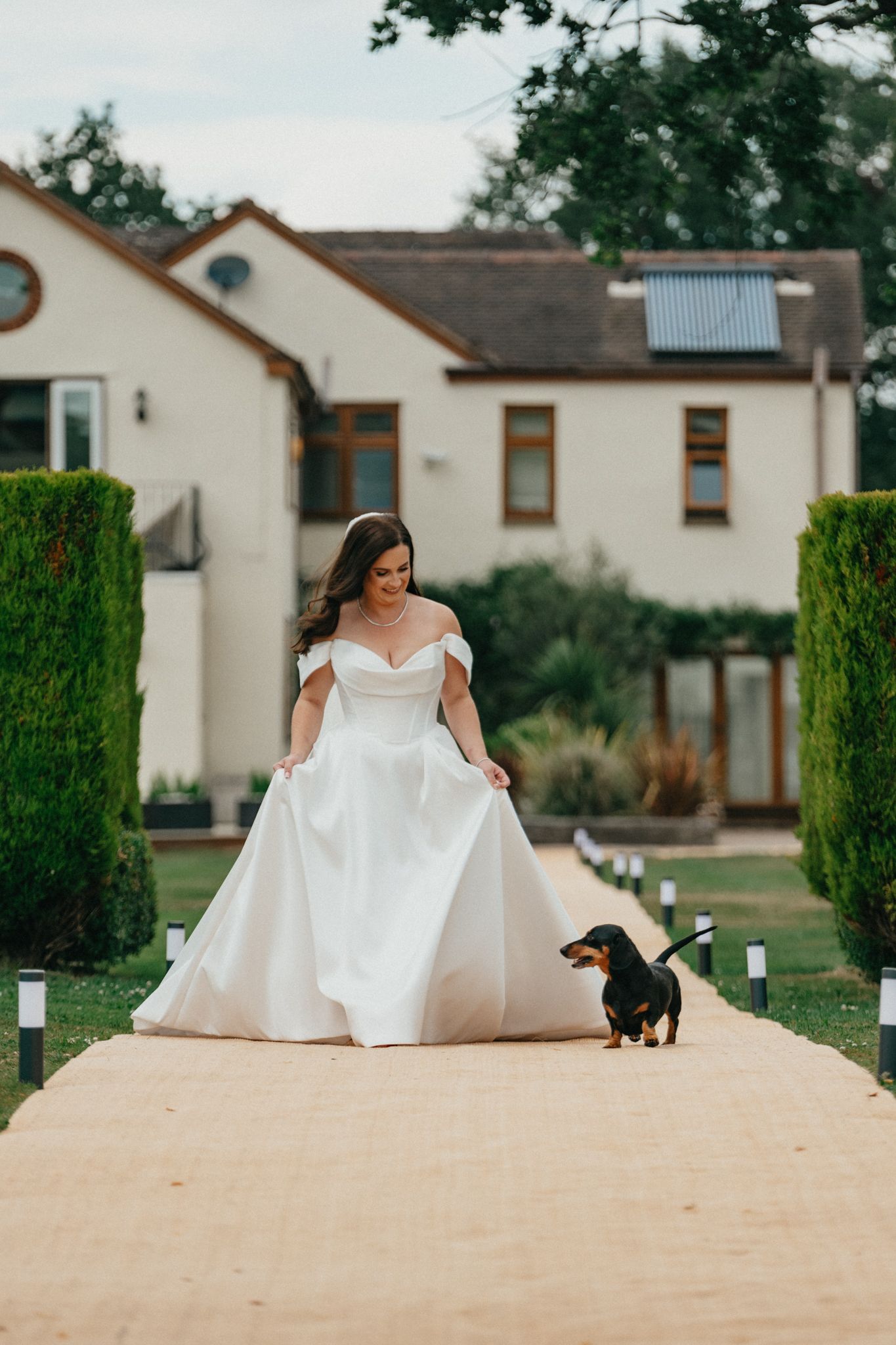 A bride in a white gown walks along a path with a small dachshund dog beside her.