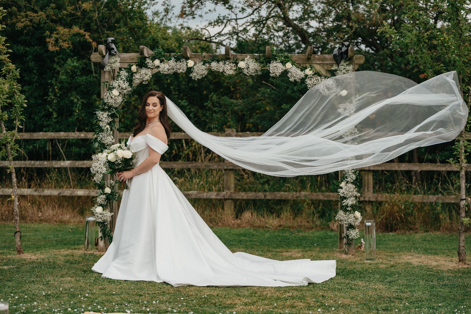Bridesmaids in navy dresses hold a flowing white wedding veil in an outdoor garden setting.