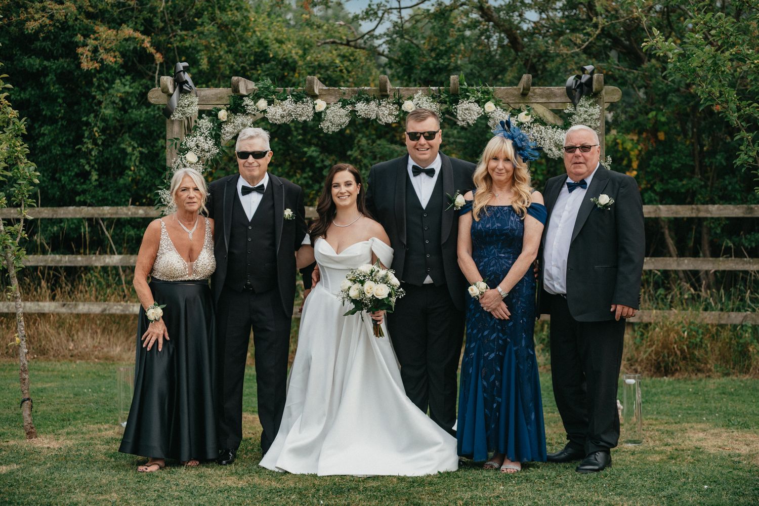 Wedding party members in formal attire pose together outdoors with a wooden fence backdrop.