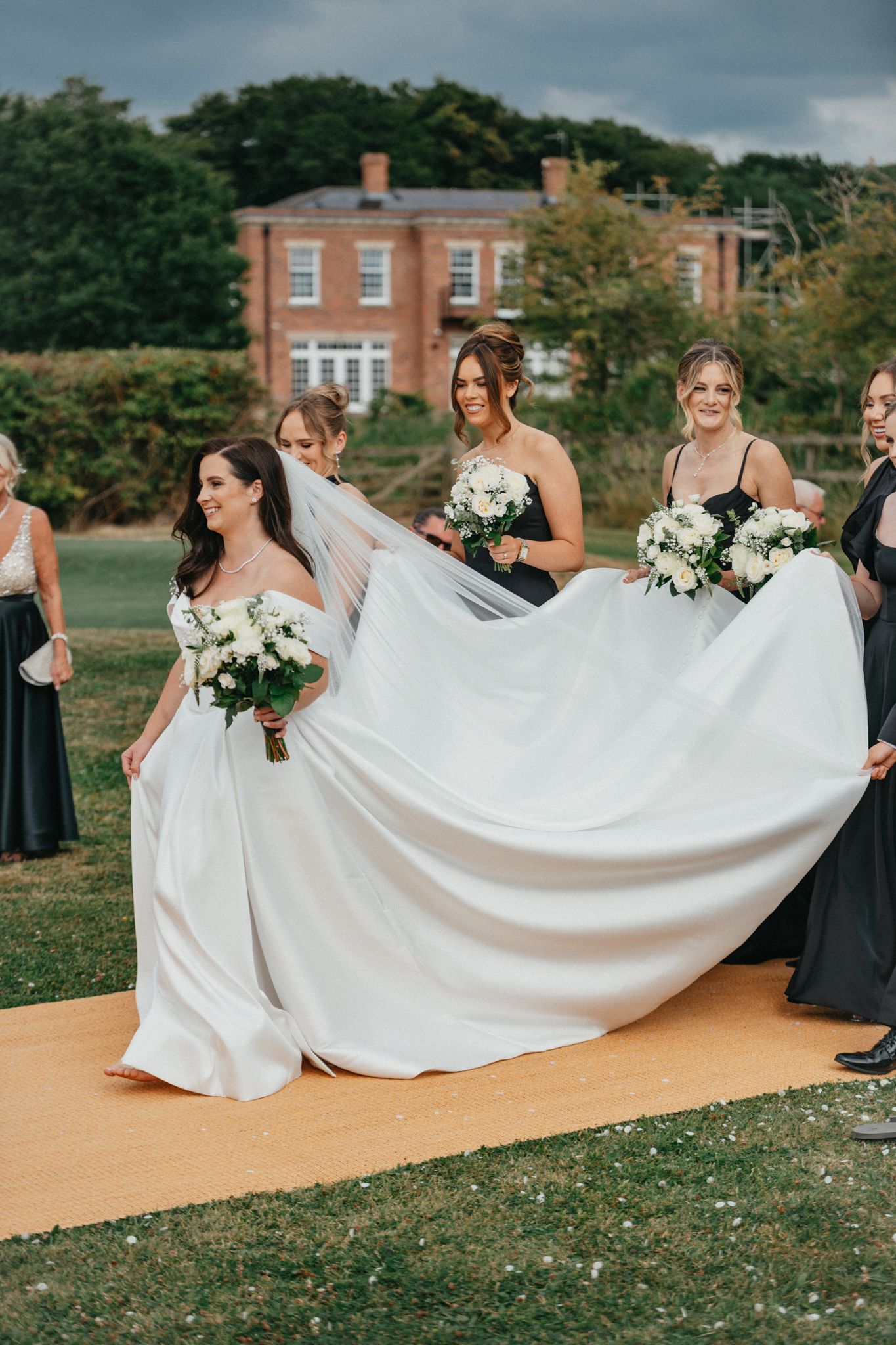Bridesmaids help arrange a long white wedding dress train on a pathway.