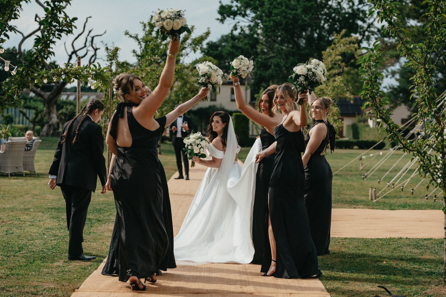 Wedding party members assist with arranging a bridal gown train outdoors.