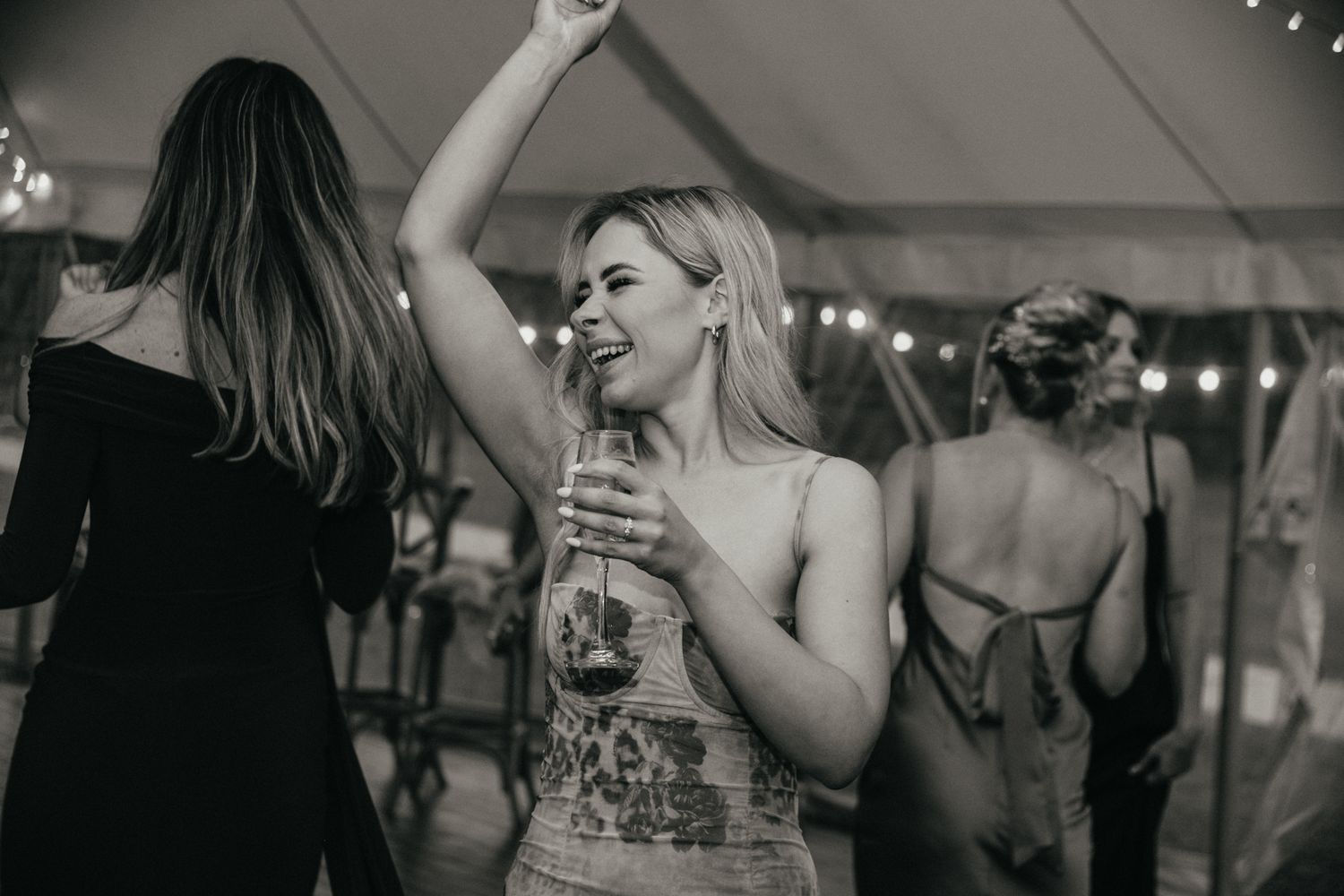 Black and white photo of reception guests dancing under tent with string lights.