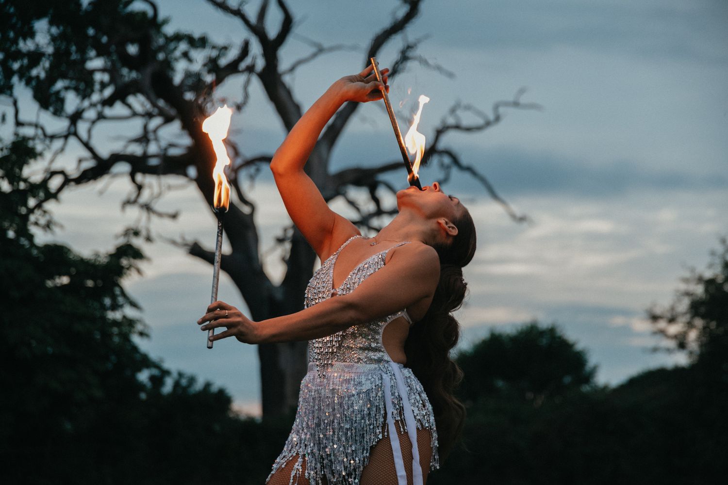 Woman in sparkly dress performs with sparklers at outdoor evening event.
