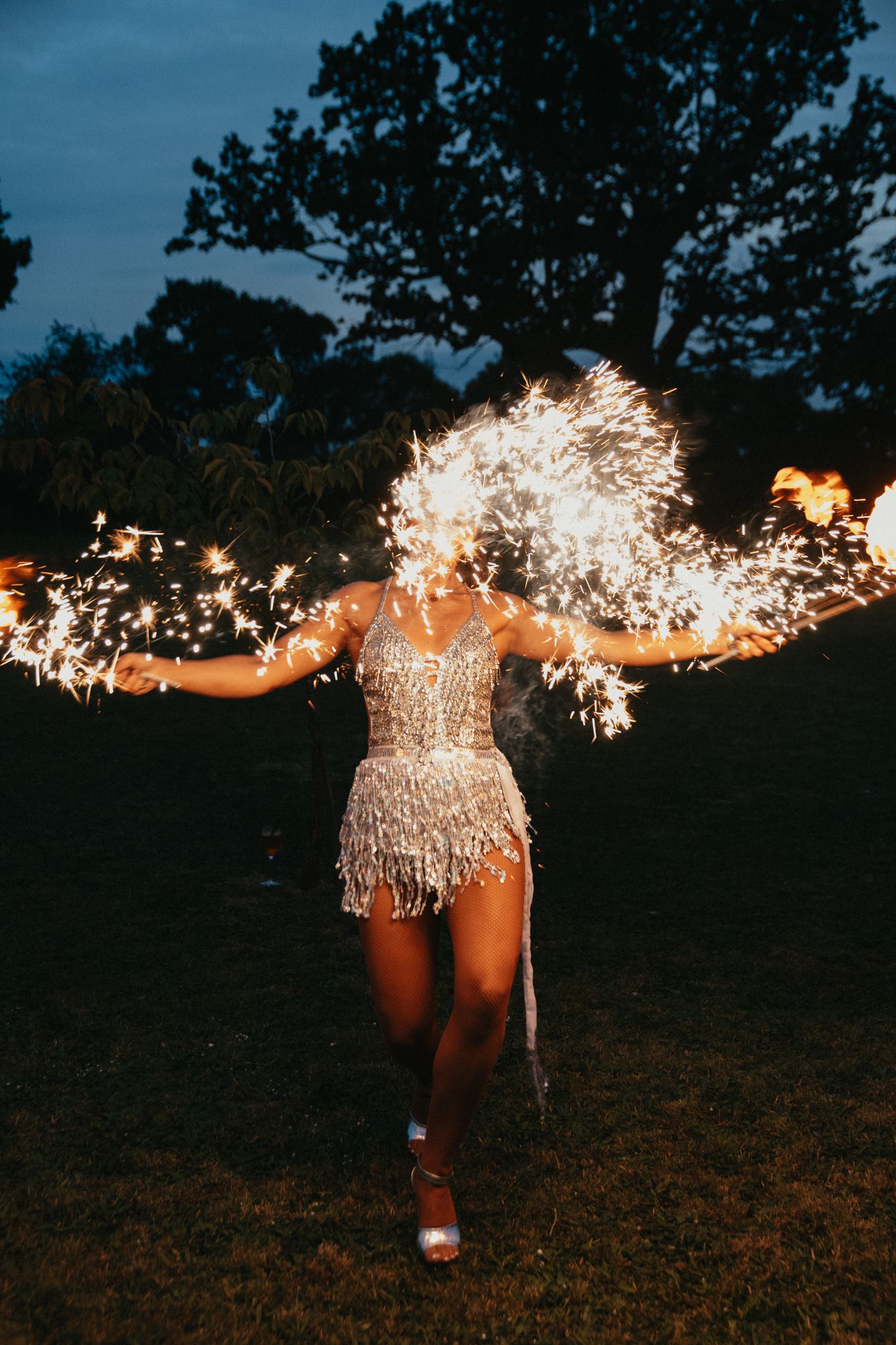 Person in a sparkly dress holds sparklers while dancing on grass at twilight during a celebration.
