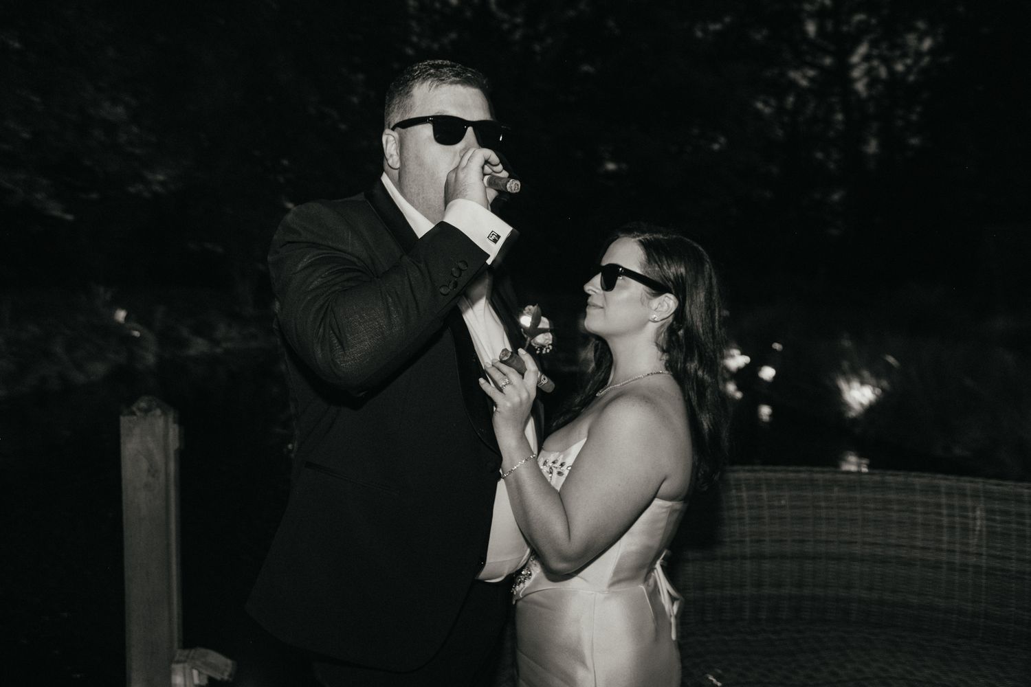 A couple wearing sunglasses share drinks outdoors at nighttime in a black and white photo.