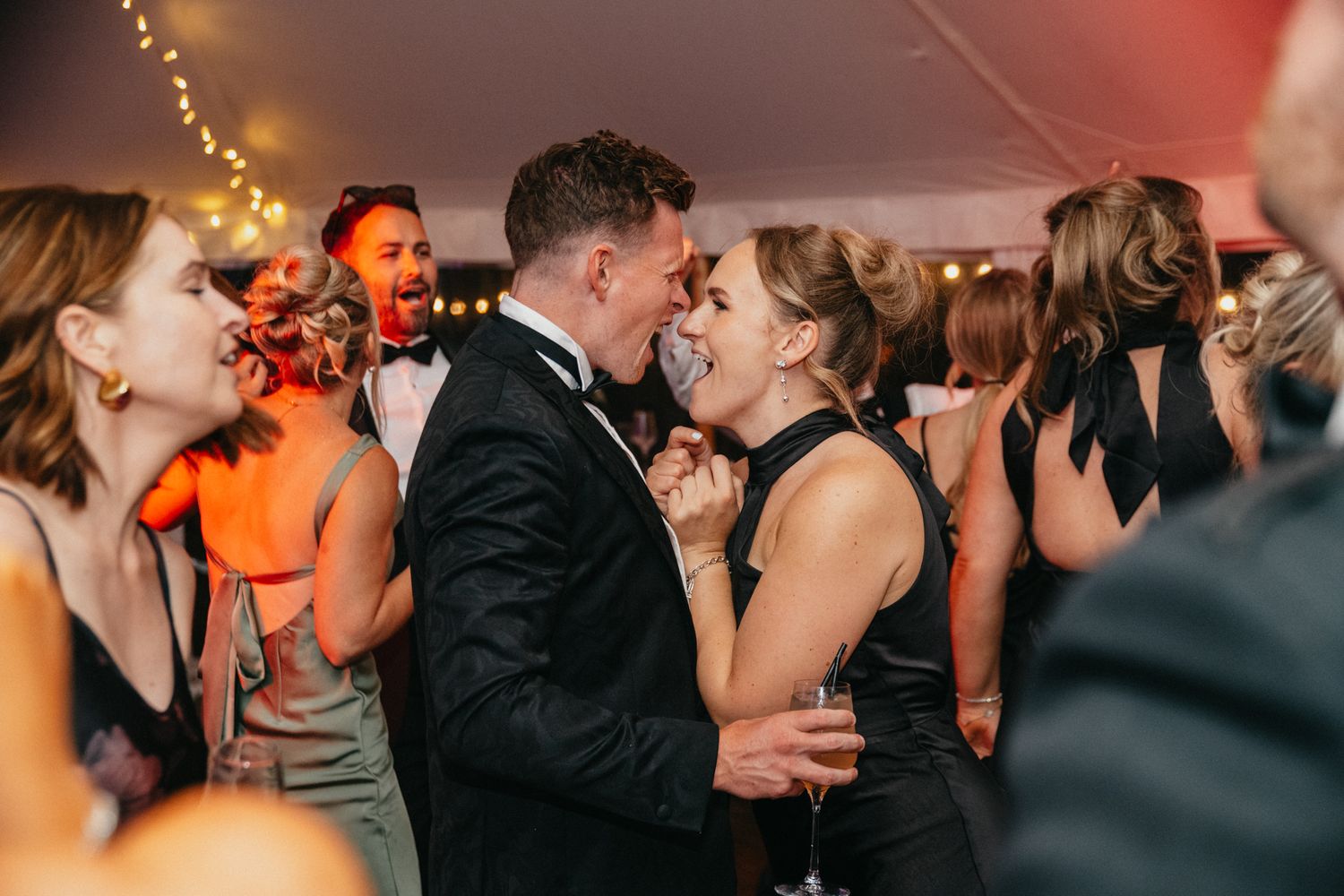 Lively crowd dancing and celebrating at a wedding reception under warm tent lighting.