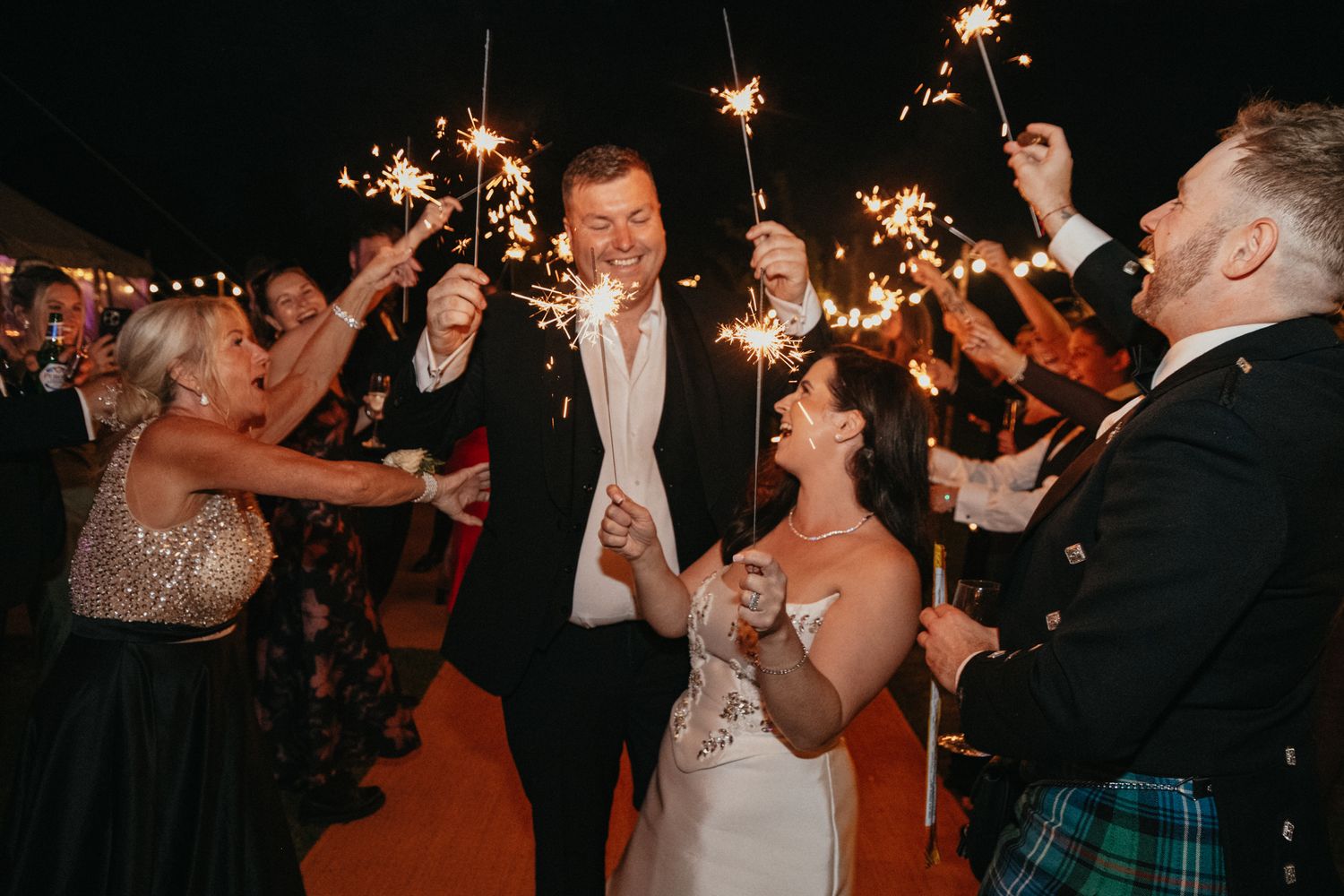 Wedding guests hold sparklers in celebration at a nighttime outdoor reception.